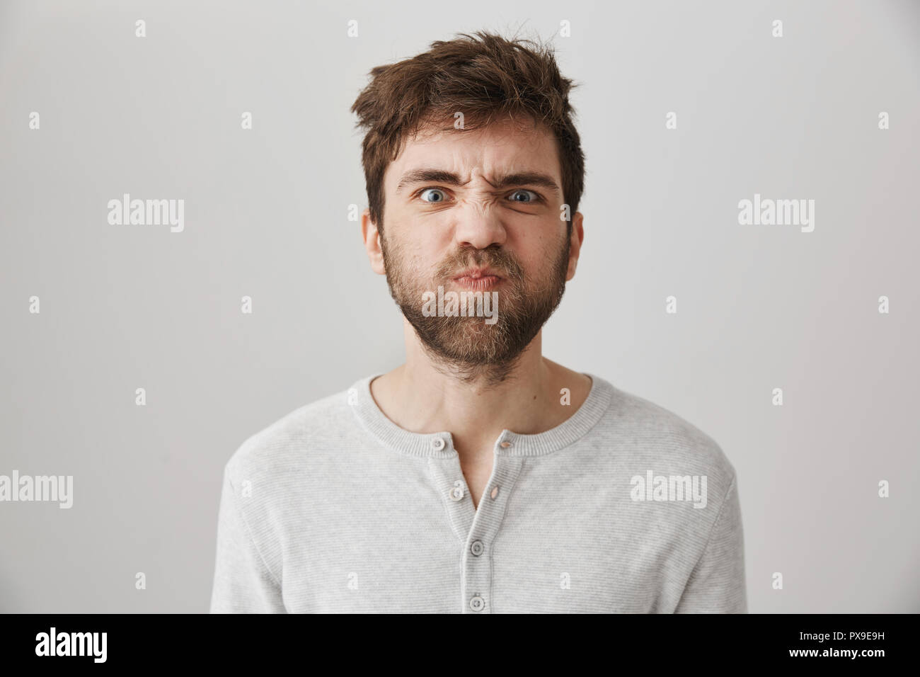 Portrait of funny weird guy with messy hair and beard making faces, puckering eyebrows and sulking, standing over gray background with offended or irritated expression, losing his temper Stock Photo