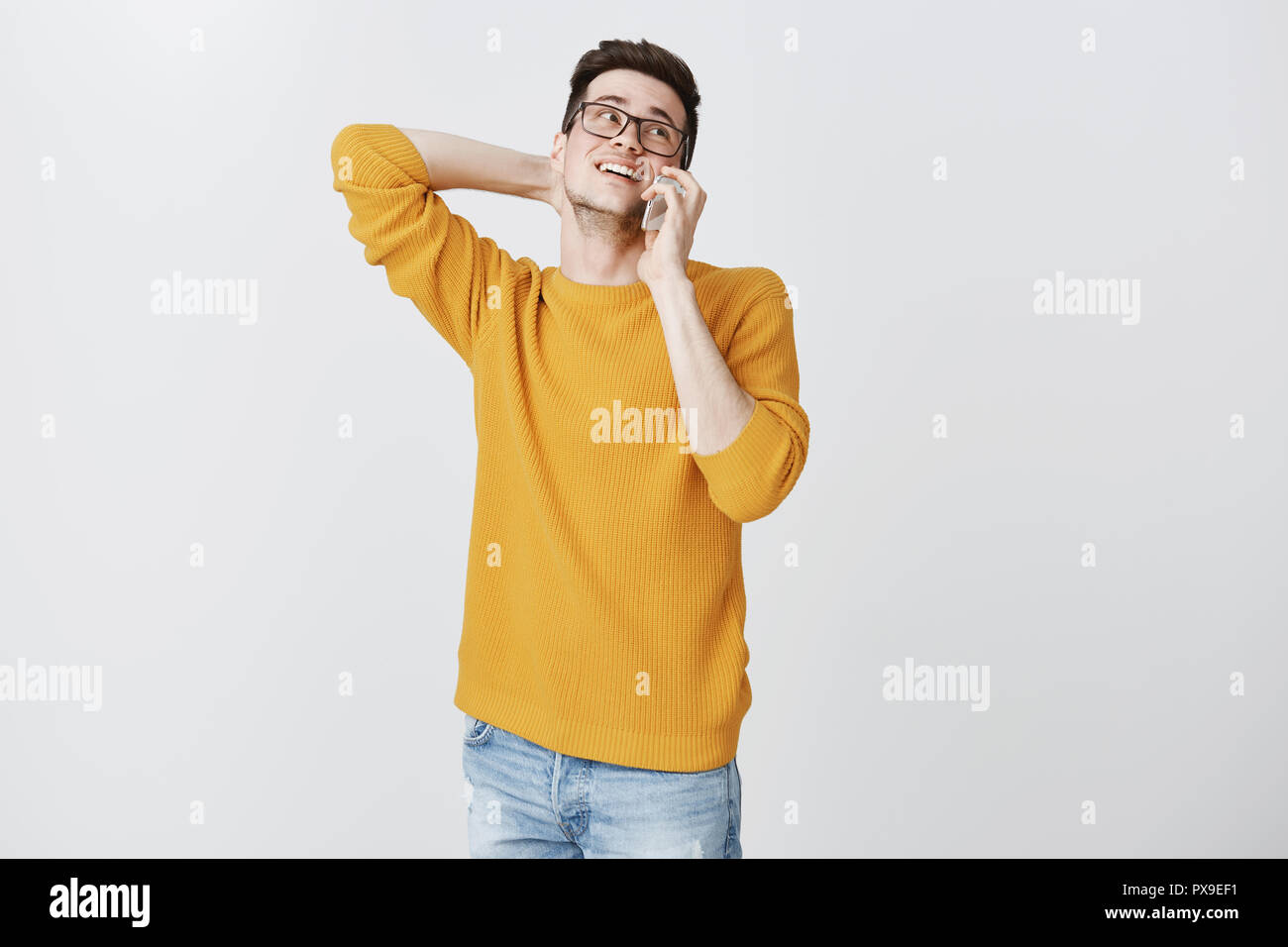 Handsome cute boyfriend being shy and hesitating scratching back of neck tilting head up as talking on smartphone trying ask out girlfriend for date smiling silly posing in sweater over grey wall Stock Photo