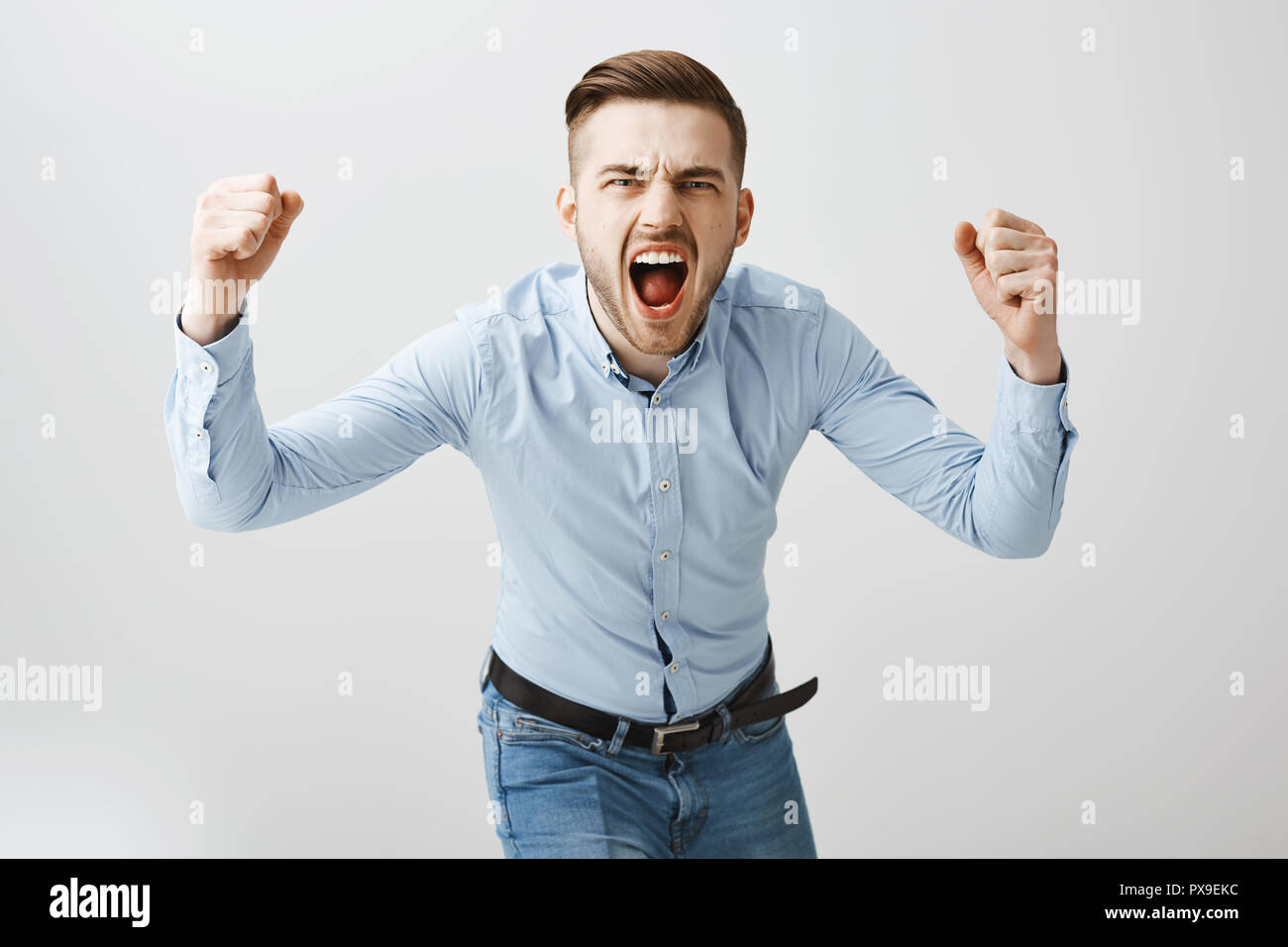 Emotive thrilled concerned european male boss in formal blue shirt and jeans bending towards camera yelling and raising fists cheering for football team making bet being angry over gray wall Stock Photo