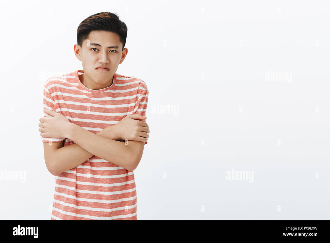 Why so cold like in fridge. Displeased intense asian teenage guy in striped t-shirt trembling hugging himself with hands crossed against chest, pursing lips, have not prepared to chilly windy weather Stock Photo