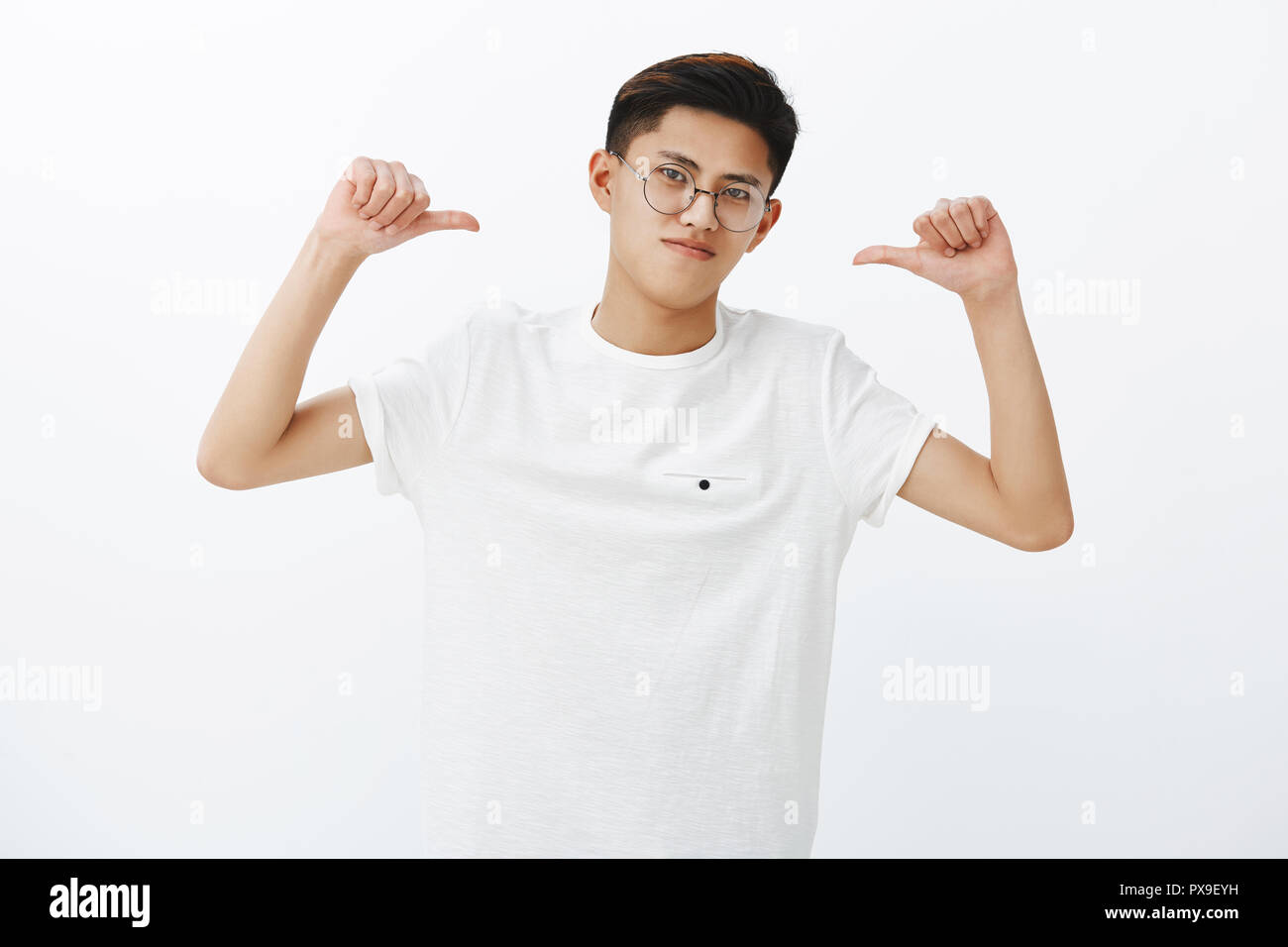Asian guy is pro feeling proud and self-assured triumphing, being winner pointing at himself with thumbs tilting head looking confident and assertive in own skills, posing in glasses and white t-shirt Stock Photo