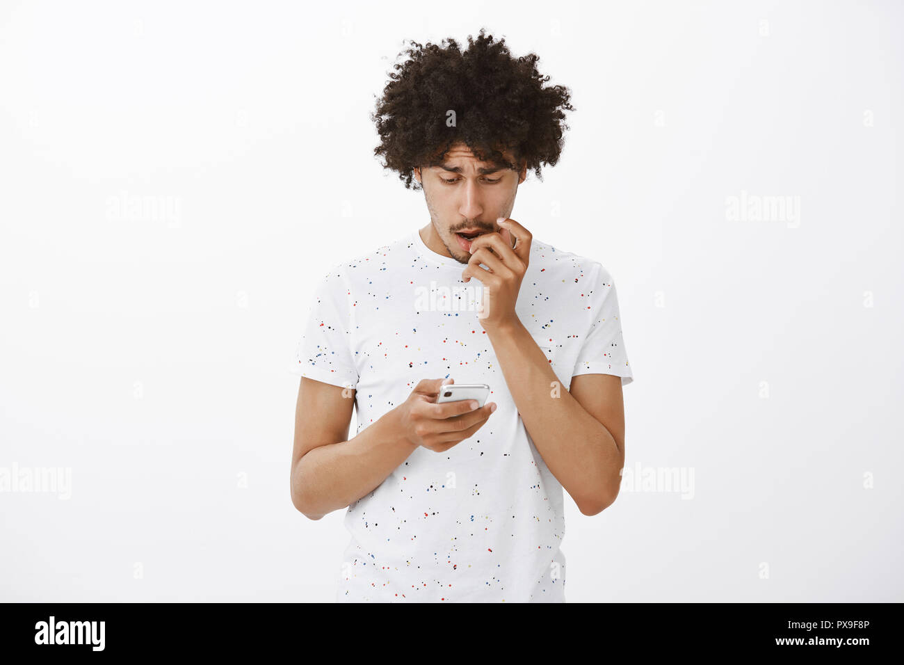 Cute hispanic football fan checking results of match via smarpthone, frowning and biting fingernail while looking worried at phone screen, losing bet, standing nervous over gray background Stock Photo