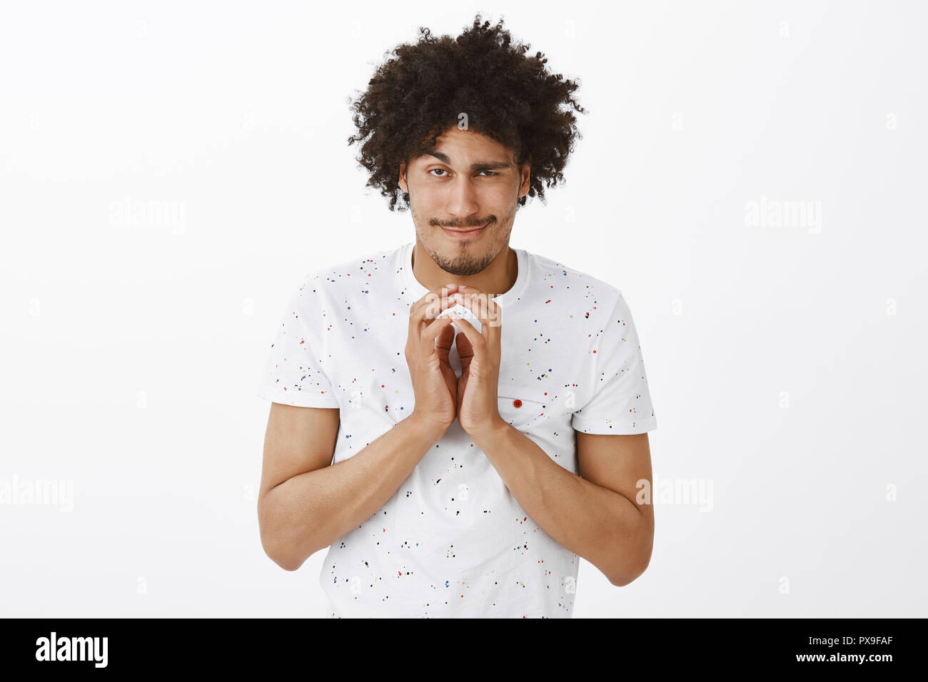 Evil genius have great plan to conquer world. Tricky and mysterious funny hispanic guy with curly hair, steeping fingers and smirking at camera, lifting one eyebrow, having bad intention in mind Stock Photo