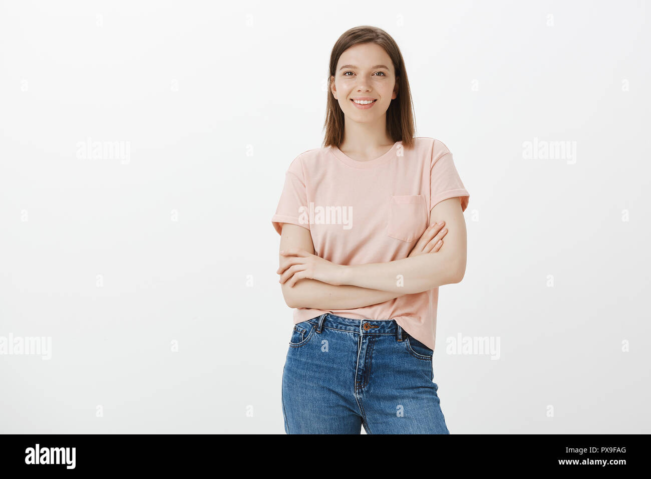 Good-looking self-assured caucasian female model in trendy t-shirt and jeans holding hands crossed on chest and smiling joyfully, expressing confident and happy attitude, standing over gray background Stock Photo