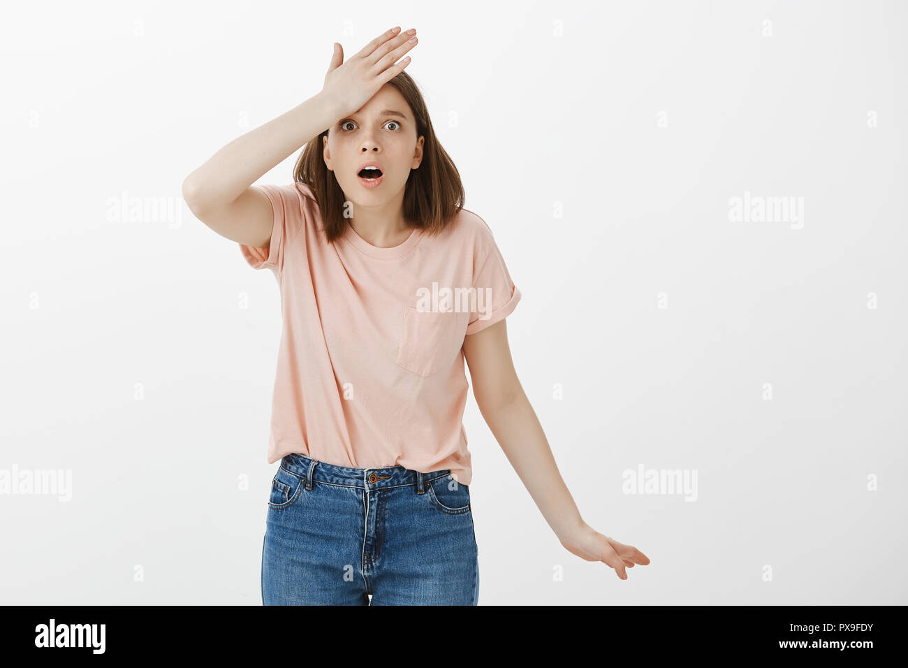 Girl remembered importrant task she forgot, punching herself in forehead, dropping jaw and standing anxious over gray background, gesturing, being forgetful and intense, recalling something Stock Photo