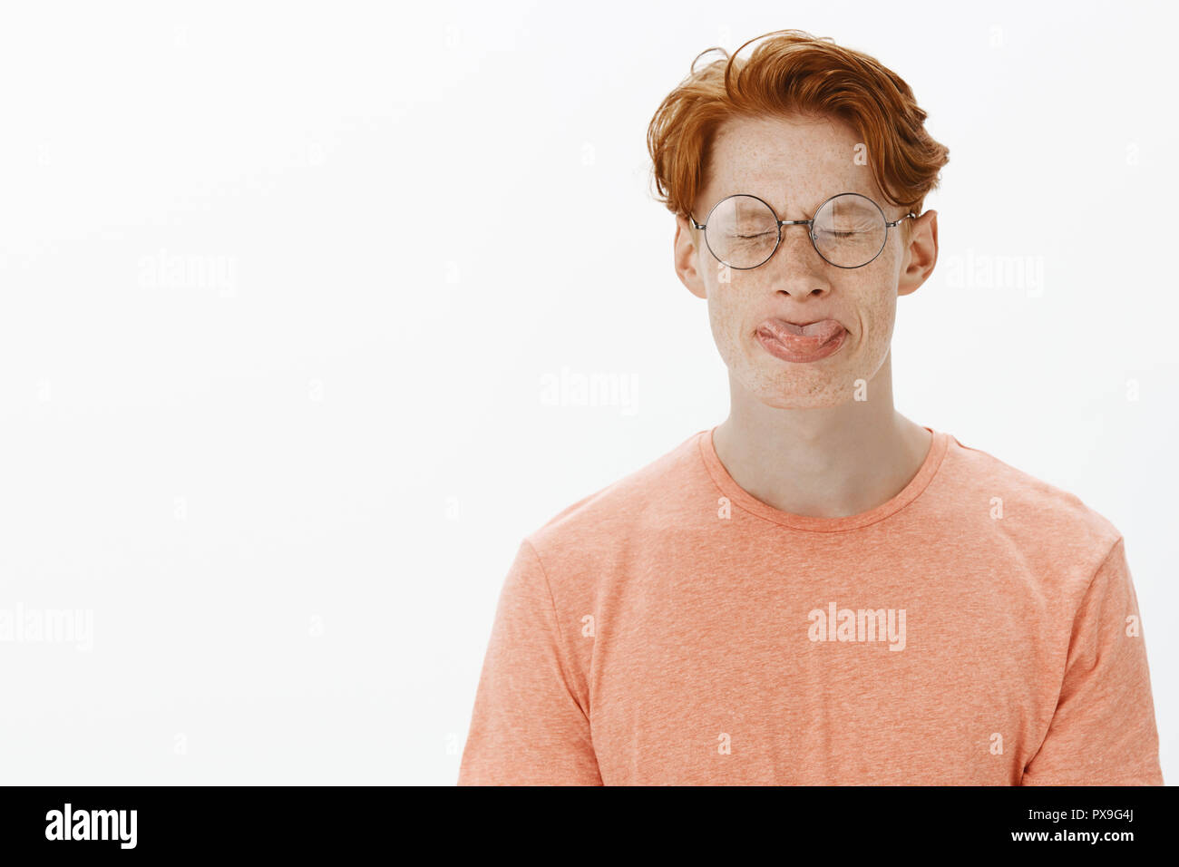 Cute and funny redhead man with freckles and stylish haircut in round glasses sticking out tongue, trying to roll it with closed eyes, fooling around, losing bet with girlfriend over gray background Stock Photo