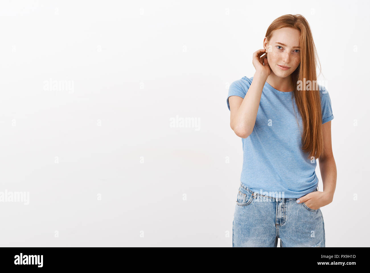 Cute young european redhead female student with freckles flicking hair behind ear holding hand in pocket and gazing with shy and awkward expression at camera asking girlfriend for date over gray wall Stock Photo