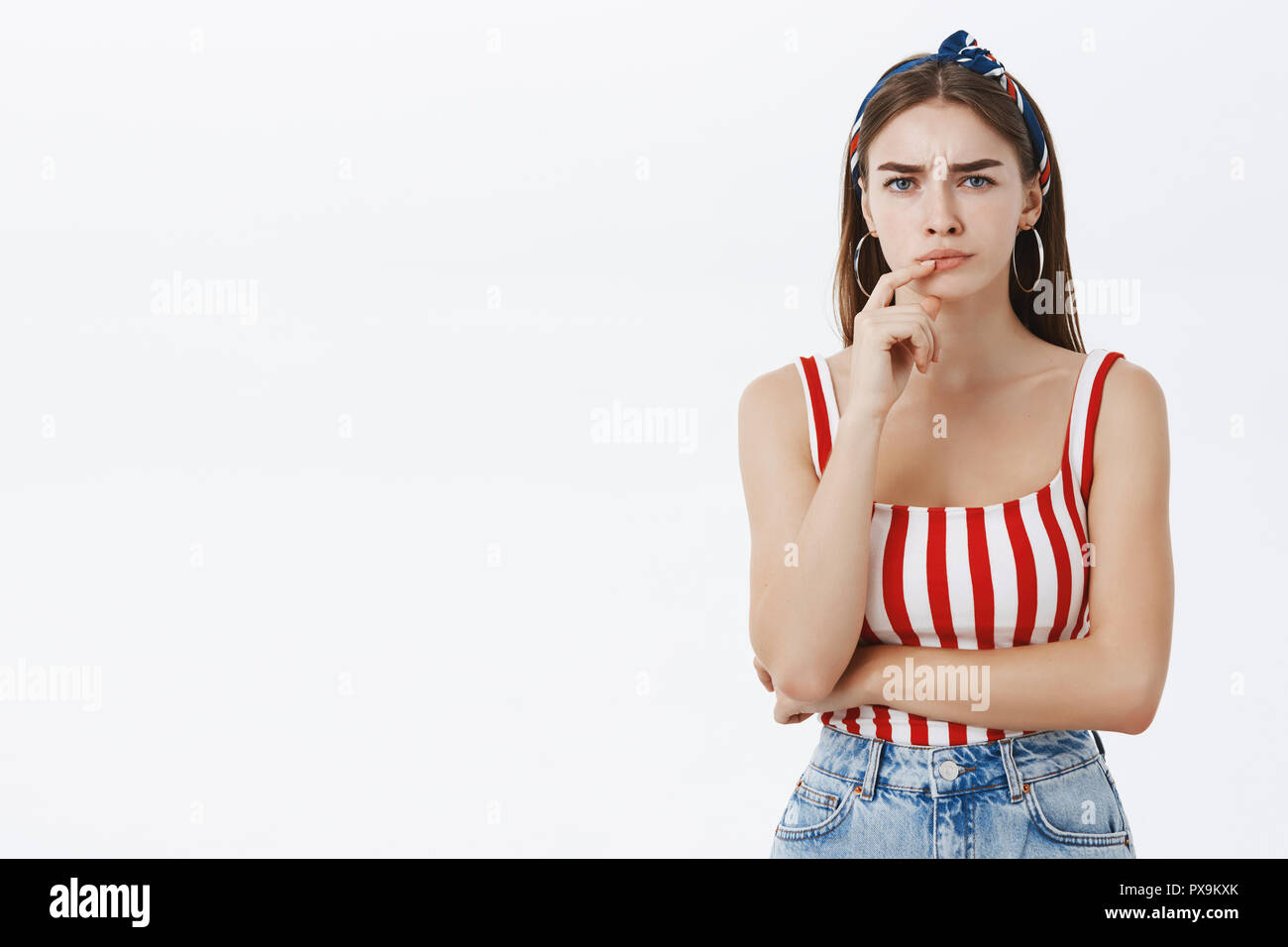 Waist-up shot of suspicious bossy woman with brown hair in striped top pursing lips holding finger on closed mouth frowning doubtfully being unsure and displeased with employee project over grey wall Stock Photo
