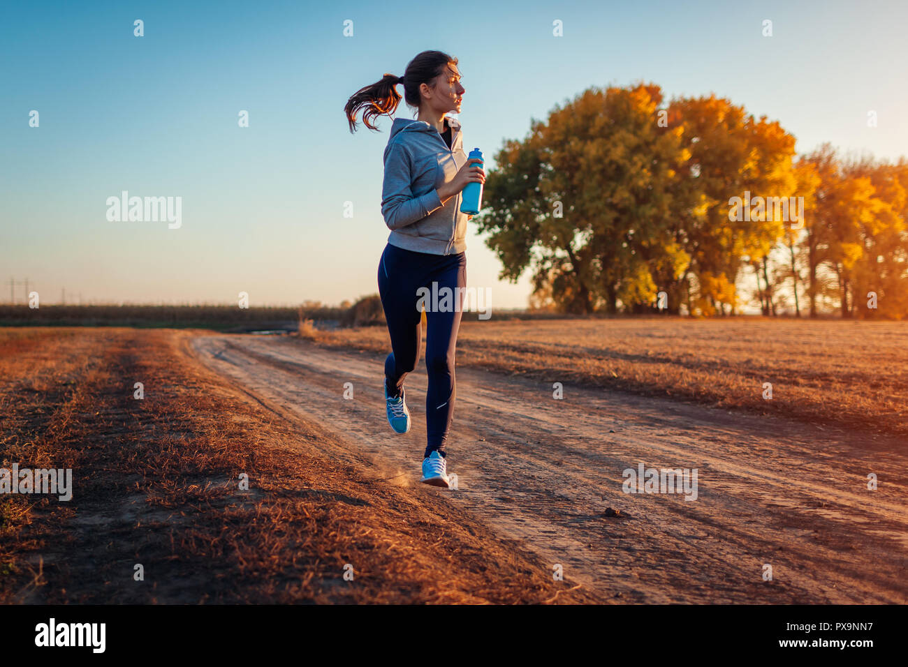 Woman running holding bottle of water in autumn field at sunset. Healthy lifestyle concept. Active sportive people Stock Photo