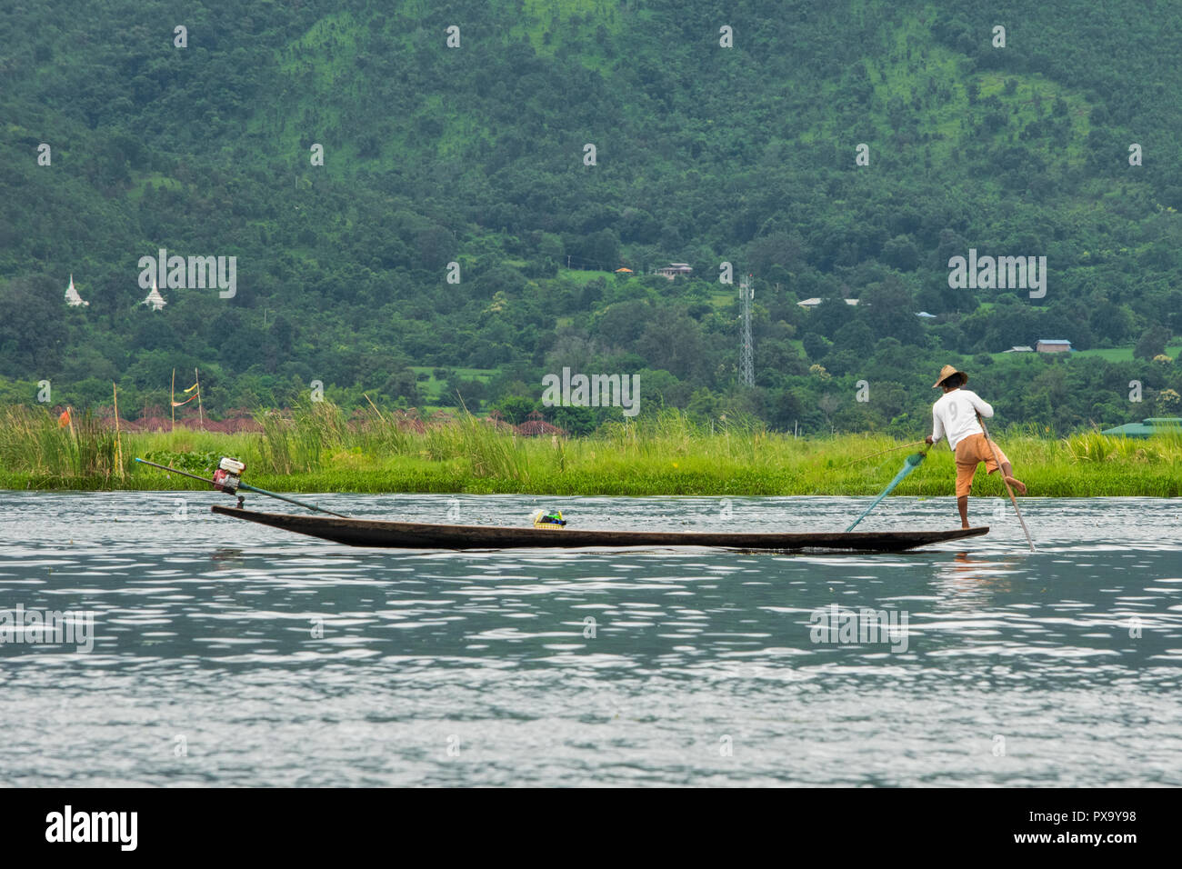 Travel, local fisherman in white shirt and orange shorts balancing on one foot on the tip of the boat and pulling net to collect fish, Inle Lake Burma Stock Photo