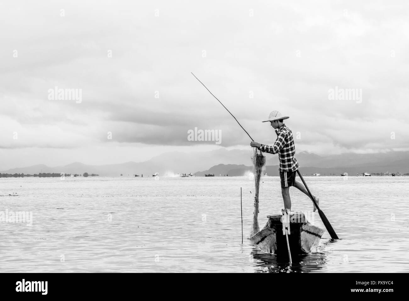 Travel local young Burmese male fisherman wearing checked shirt, using stick and net to fish, balancing on one foot on boat, Inle Lake Myanmar, Burma Stock Photo