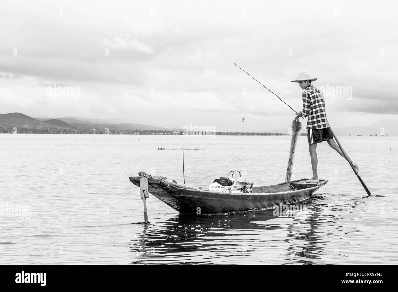 Travel local young Burmese male fisherman wearing checked shirt, using stick and net to fish, balancing on one foot on boat, Inle Lake Myanmar, Burma Stock Photo