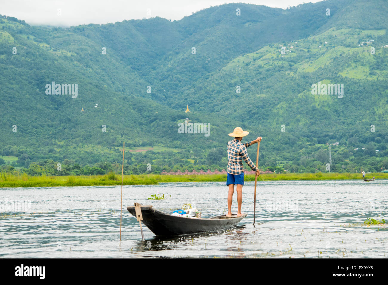 Local young Burmese fisherman wearing a checked shirt returning home after fishing, standing on the edge of a thin tall boat, rowing back, Inle Lake Stock Photo