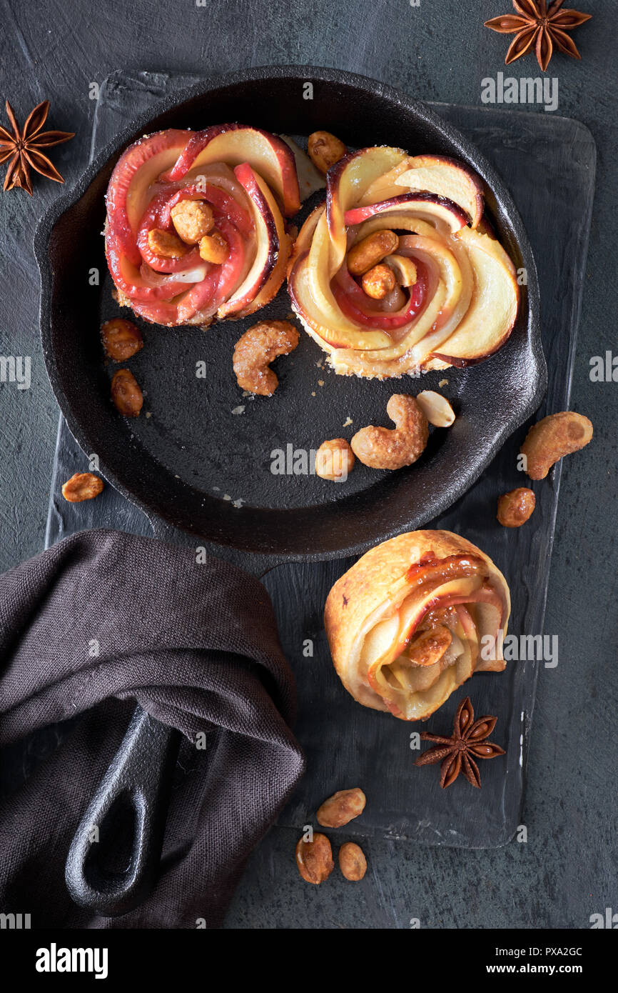 Three homemade puff pastries with rose shaped apple slices baked in cast iron skillet. Top lay on dark background with linen towel and caramelized nut Stock Photo