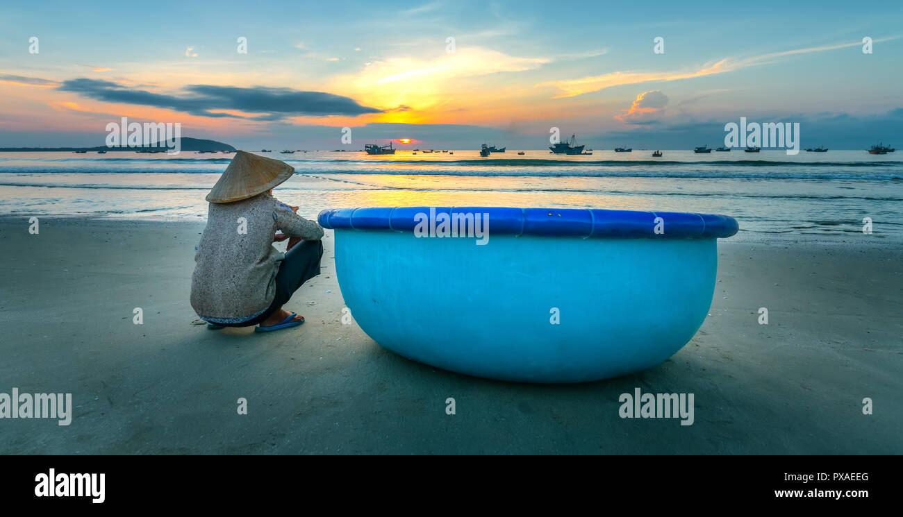 Old fisherman seated on a beautiful beach at dawn next to boat looking relatives fishing shore hoping for bumper harvest Stock Photo
