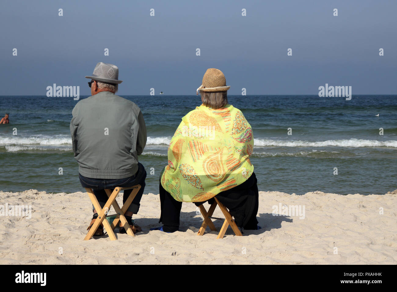 An elderly couple sit on wooden stools on the beach and watch the sea. Stock Photo