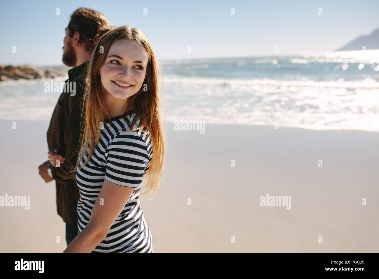 Pretty young woman looking back and smiling while walking on the beach with her boyfriend. Young couple on the sea shore. Stock Photo