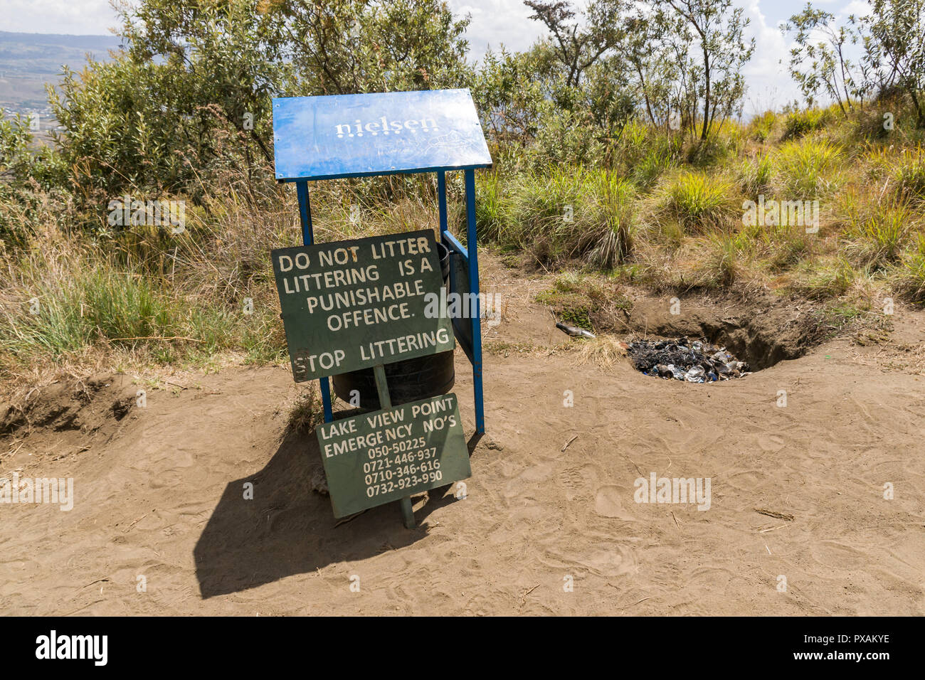 A pile of burnt rubbish lies in a small hole by a rubbish bin with warning sign to not litter on the trail up Mount Longonot, Kenya Stock Photo
