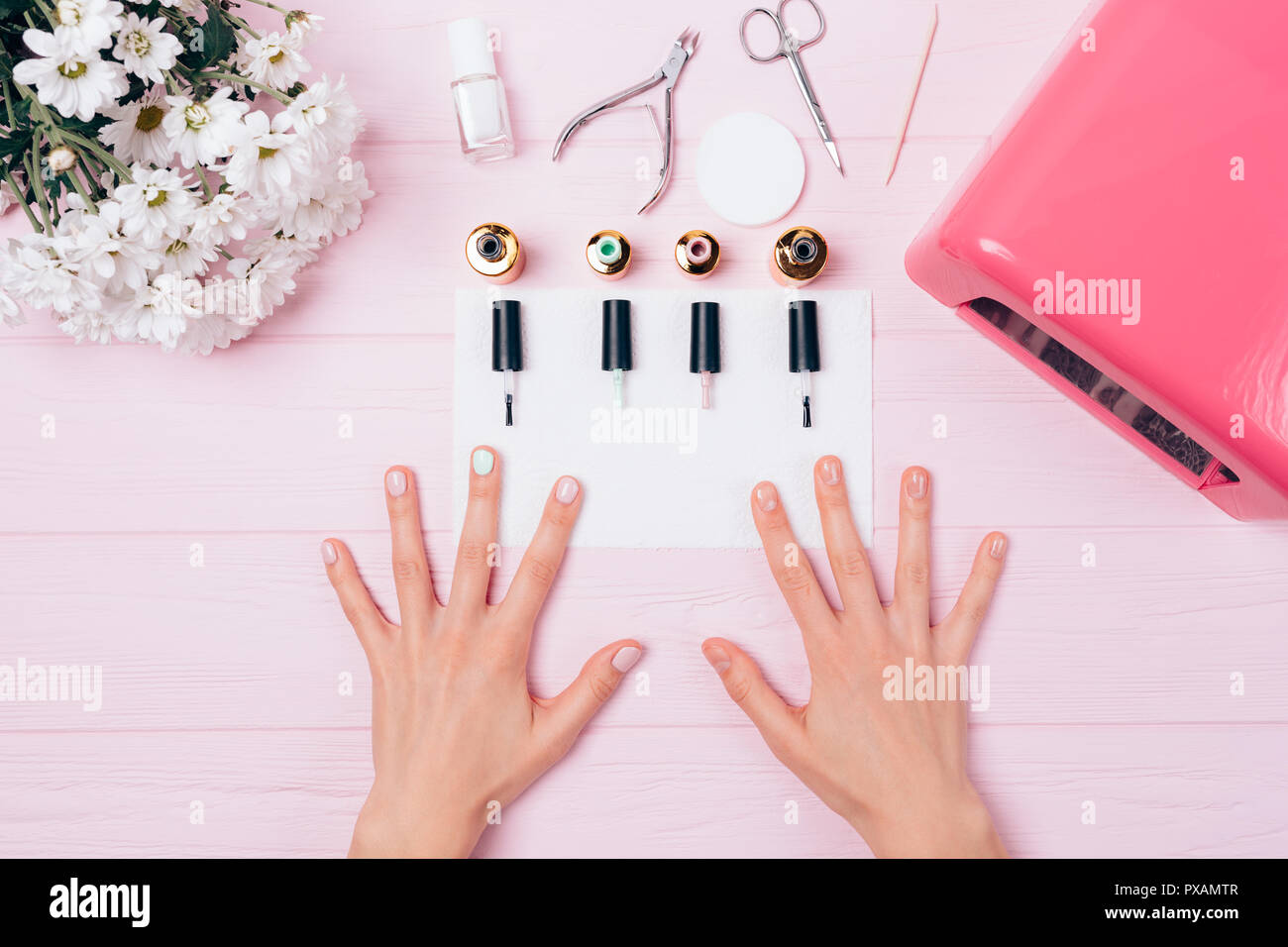 Female hands with shellac covering near opened bottles gel nail polishes, brushes, ultraviolet lamp and bouquet of flowers, top view. Creative flatlay Stock Photo