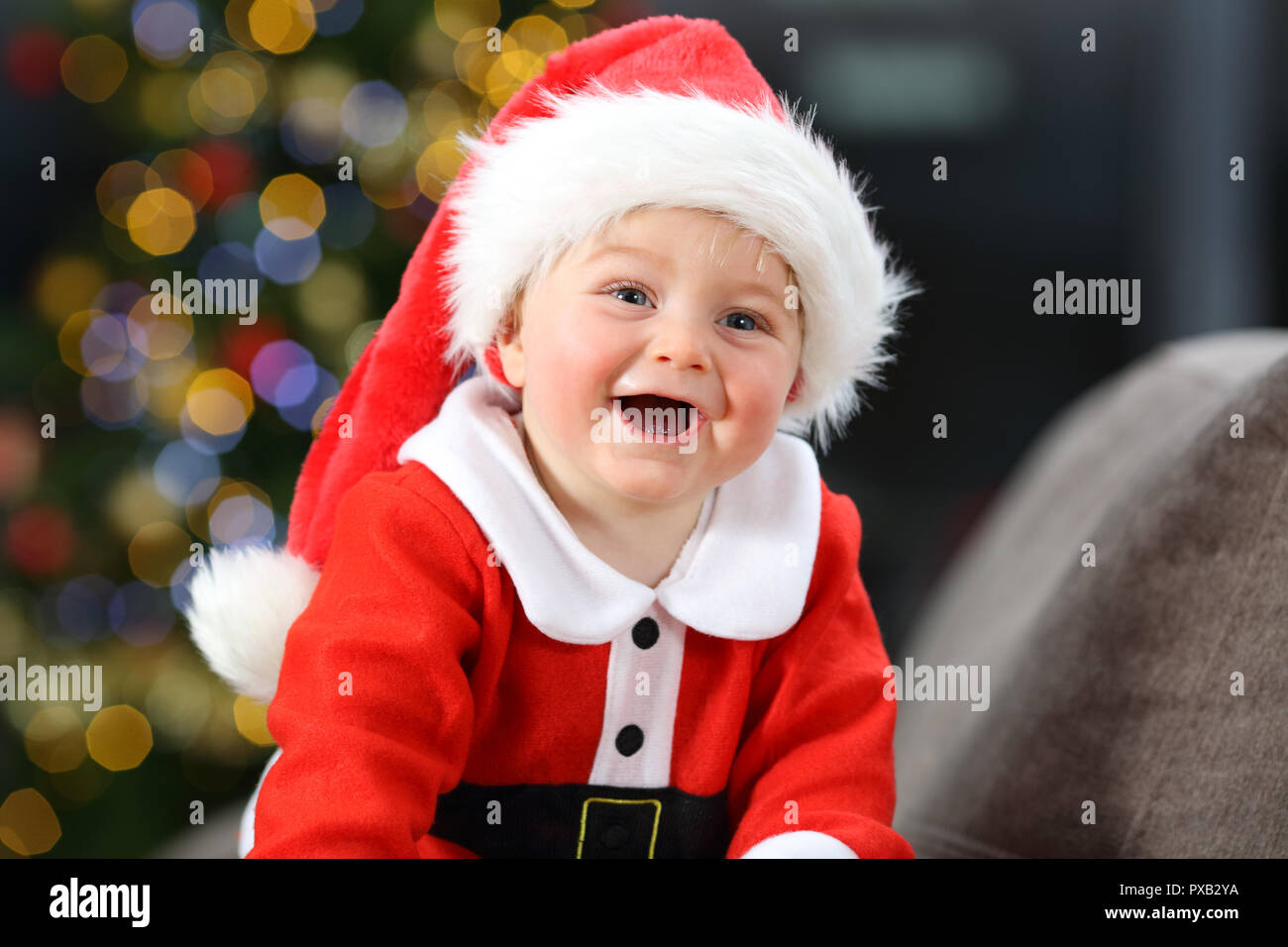 Cheerful baby dressed as santa claus on a couch at home in christmas with a tree in the background Stock Photo