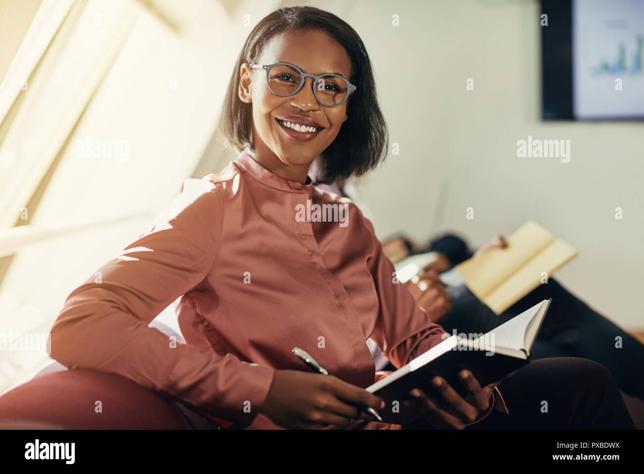 Smiling young African businesswoman writing in a notebook while sitting ...