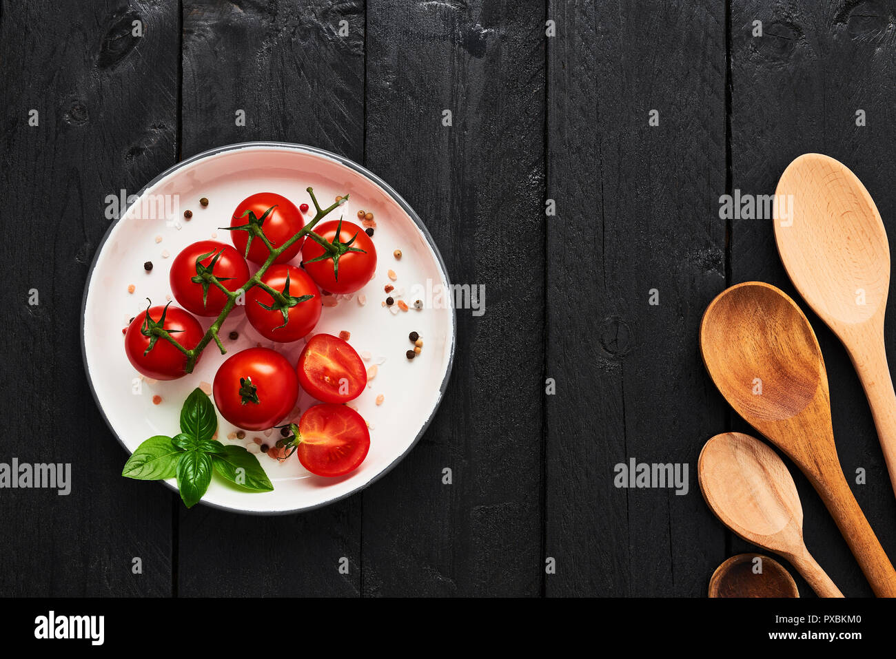 Various wooden spoons and tomatoes in a plate with Himalayan salt, pepper and basil on black wooden table. Top view with copy space for menu or recipe Stock Photo