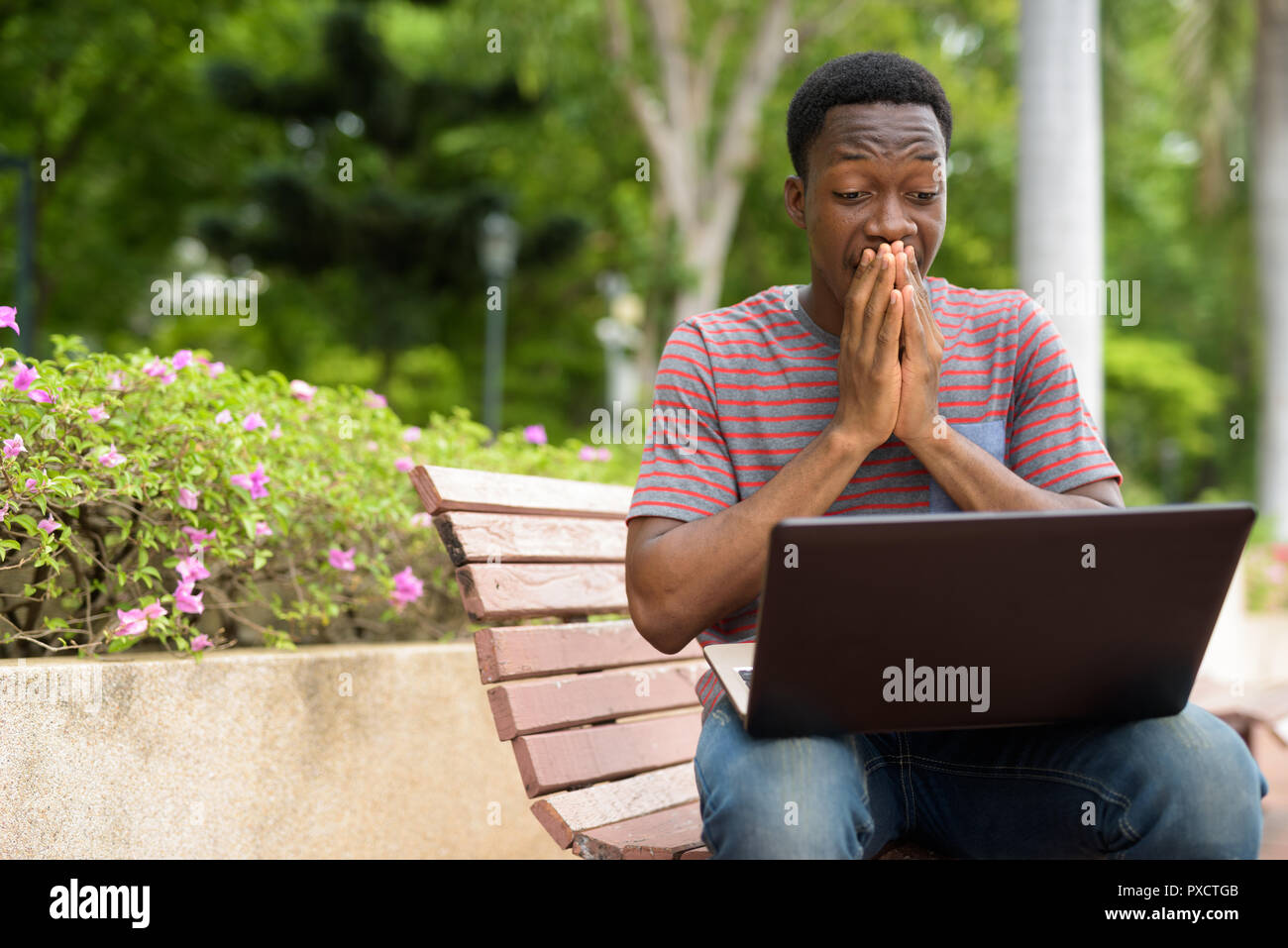 Shocked handsome African man using laptop in park while looking surprised Stock Photo