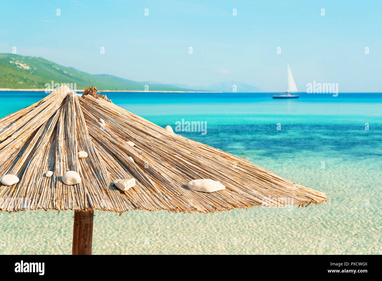 solo wooden beach umbrella on empty beach with clear waters, mountains and yacht at distance on sunny summer day at Sakarun beach, Dugi island, Croati Stock Photo