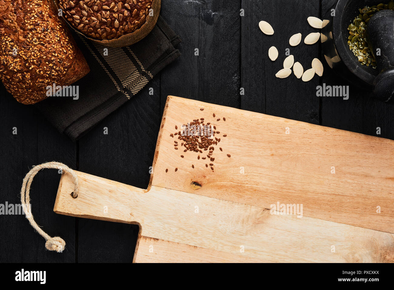 Chopping board with linseed and various crusty whole wheat bread, pumpkin seeds and mortar on black wooden table. Top view with copy space for text, m Stock Photo