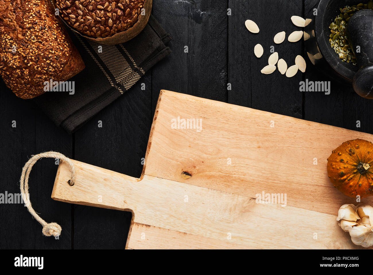 Chopping board and various crusty whole wheat bread, garlic, pumpkin seeds and mortar on black wooden table. Top view with copy space for text, menu o Stock Photo