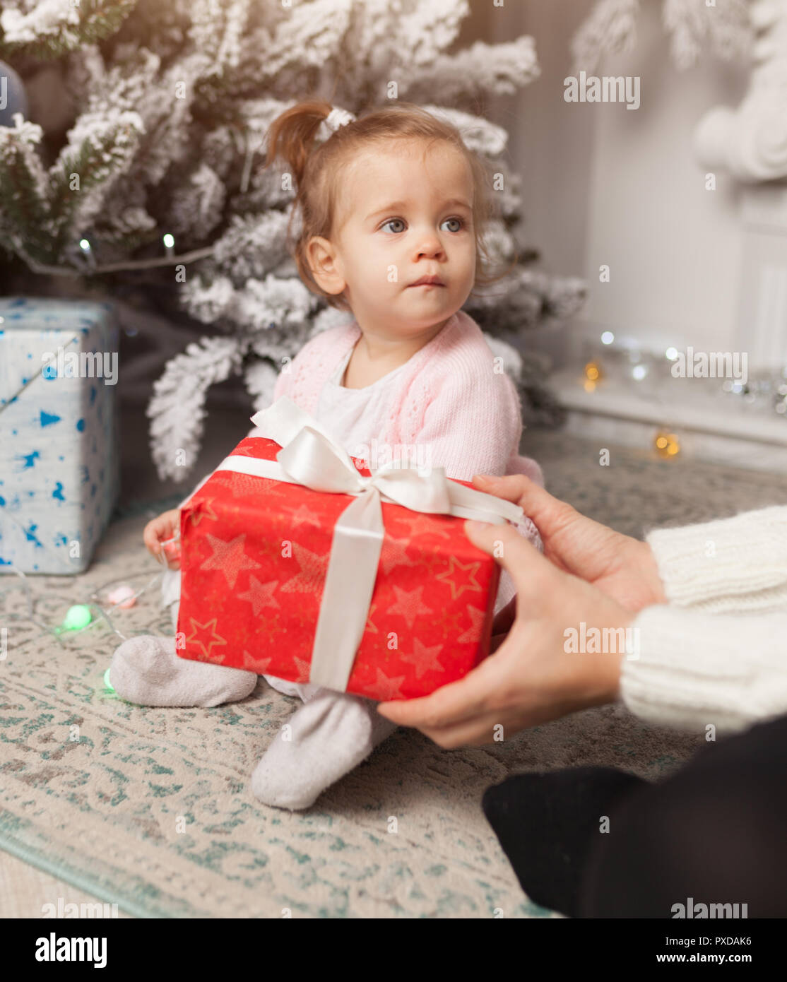 A beautiful cute little girl opens a New Year's gift. With Christmas tree on the background Merry Christmas and Happy New Year Stock Photo