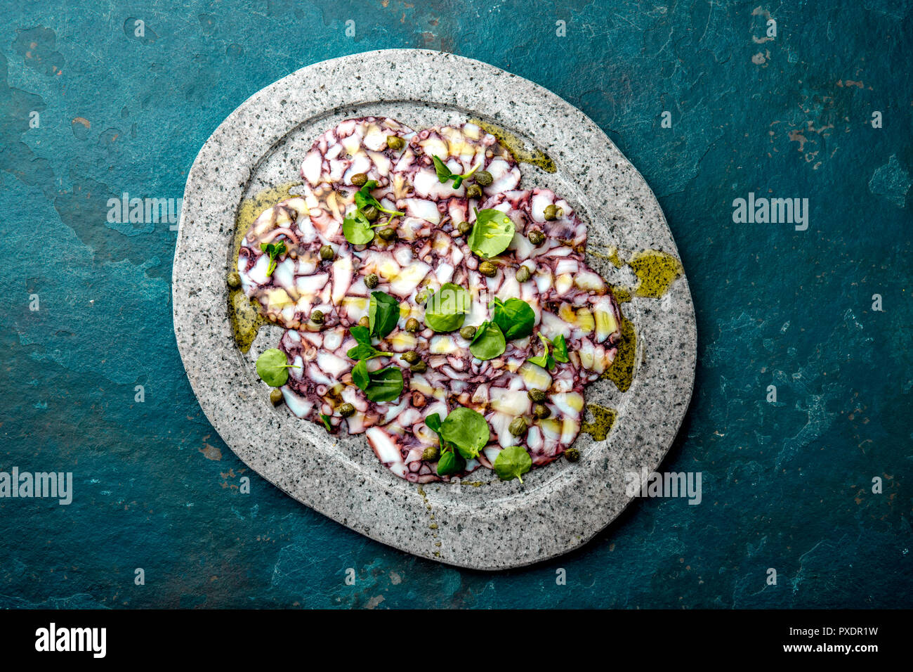 OCTOPUS CARPACCIO. Seafood Raw octopus slices with olive oil, lemon and capers on gray stone plate. Top view. blue background. Stock Photo