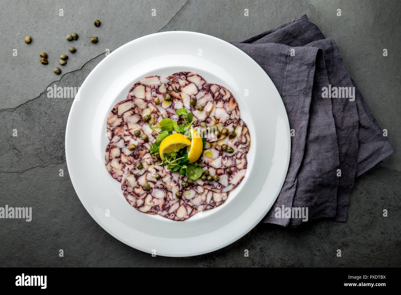 OCTOPUS CARPACCIO. Seafood Raw octopus slices with olive oil, lemon and capers on white plate. Top view. Gray stone background. Stock Photo