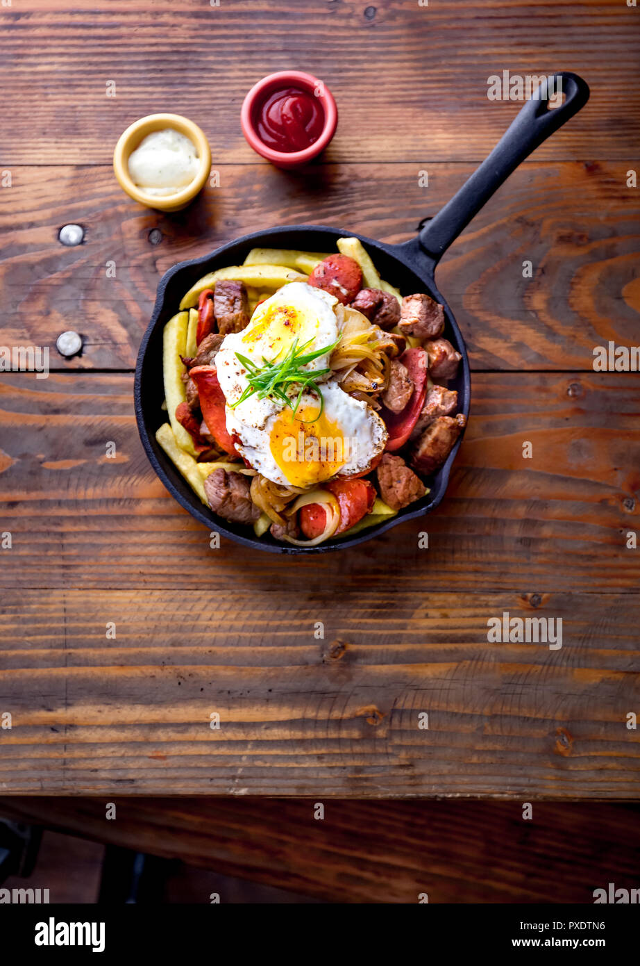 CHILEAN FOOD. CHORRILLANA - french fries topped with beef sliced, tipical sausages chorrisos, fried onion and eggs served in cast iron pan with sauces. Wooden background. top view Stock Photo