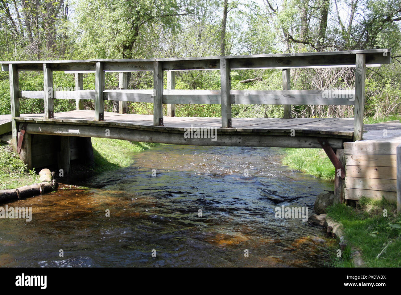 Picturesque wooden bridge spans the rushing water and rocks of a quaint village park. Eddies of pooling water bubble along green tree and grassy banks Stock Photo