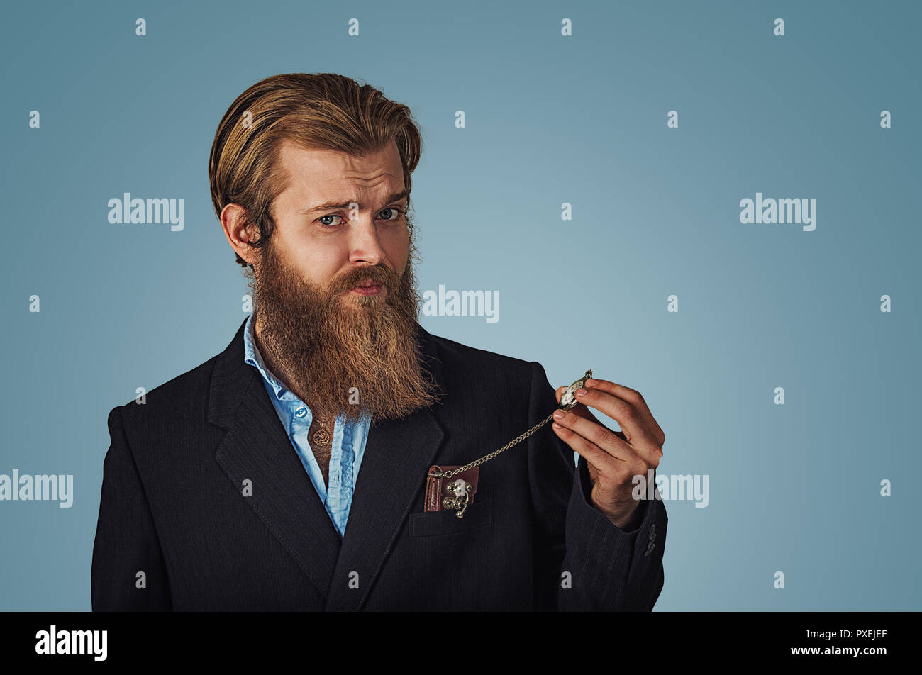 Bearded hipster Businessman looking skeptical holding retro pocket watch Isolated on blue Background. Negative face expression, human emotion, body la Stock Photo