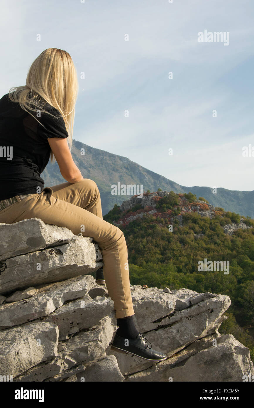 Young blond woman sitting on the edge of the mountain cliff against ...