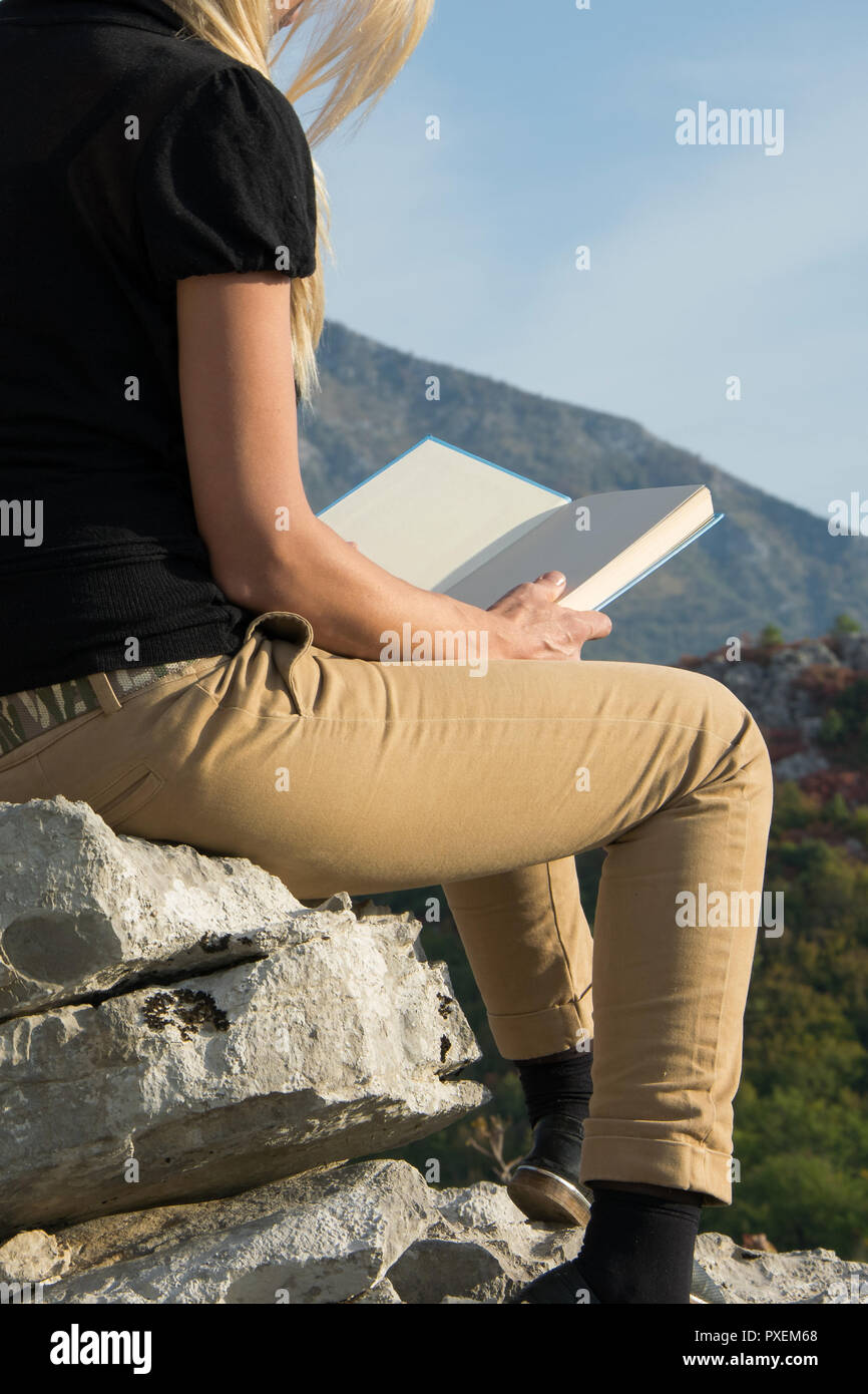 Young blond woman sitting on the edge of the mountain cliff reading a ...