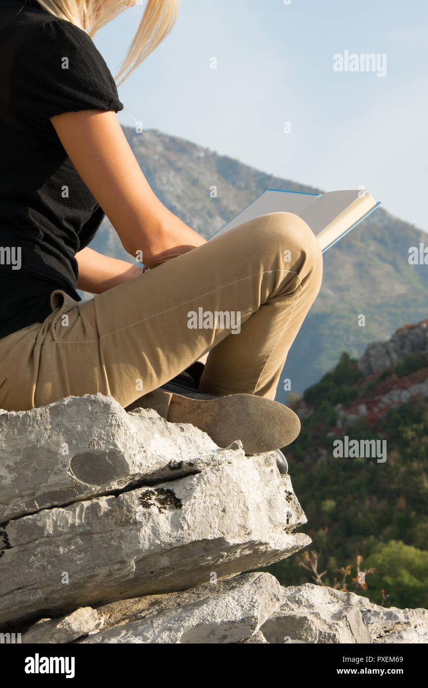 Young blond woman sitting on the edge of the mountain cliff reading a ...