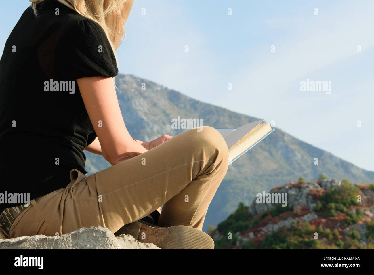 Young blond woman sitting on the edge of the mountain cliff reading a ...