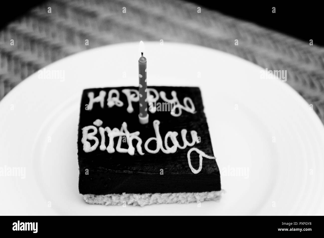 A small square chocolate birthday cake with the inscription happy birthday and one candle on a white plate. Monochrome, black and white photo Stock Photo