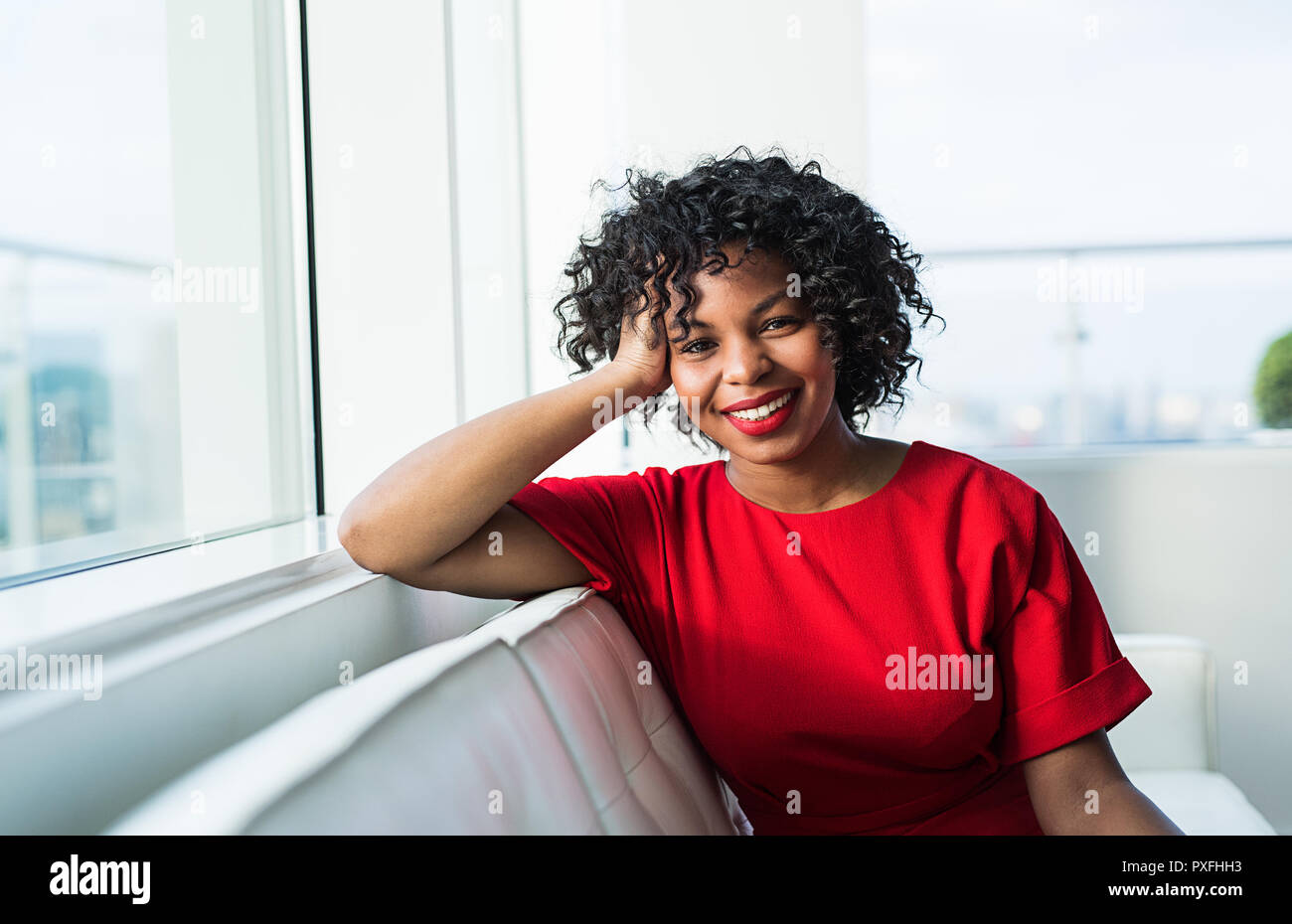 A portrait of happy black woman sitting on a sofa by the window in an ...