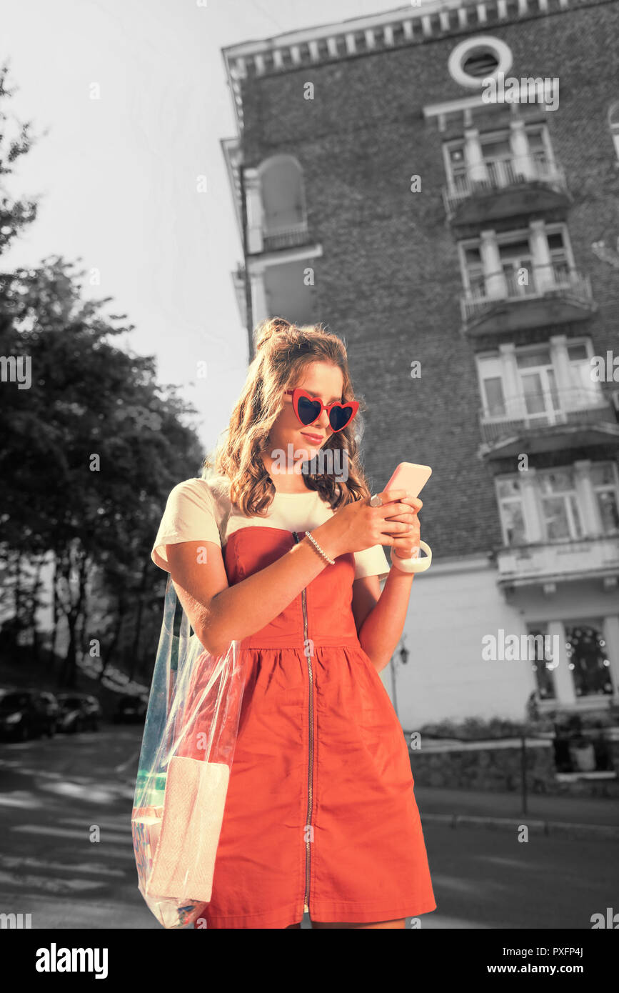 Cool stylish student wearing red dress holding pink smartphone reading messages Stock Photo