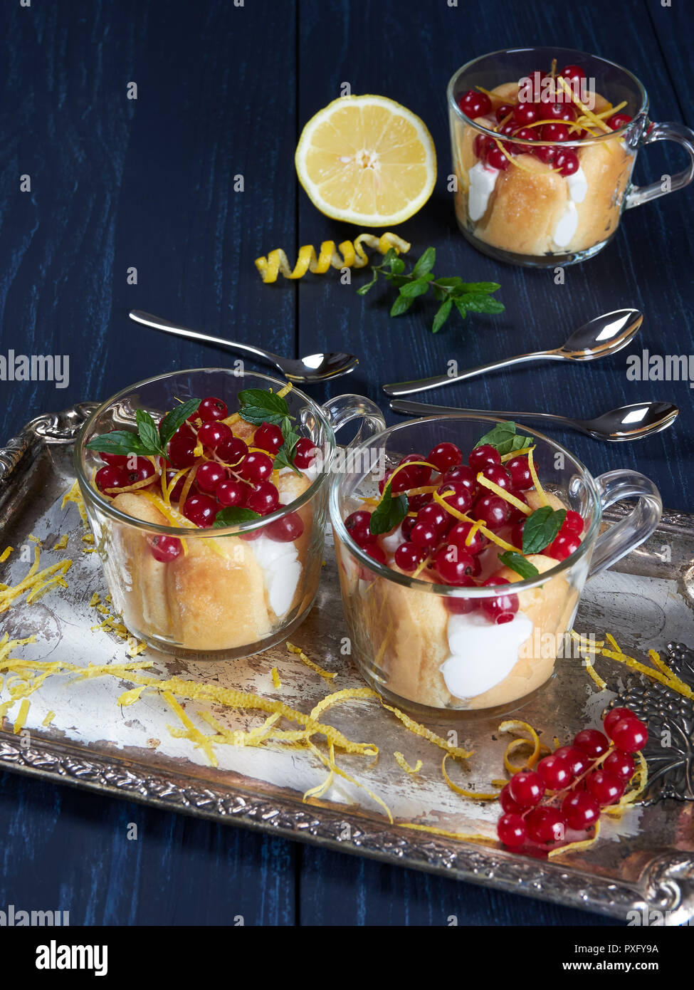 Three glass cups of limoncello (or limoncino) tiramisu topped with redcurrants and mint leaves, on a silver tray set on a dark background Stock Photo