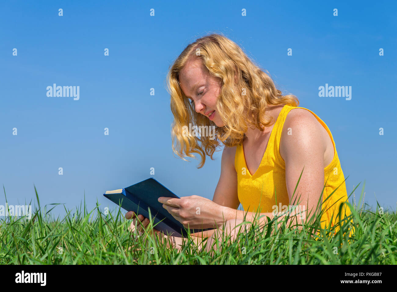 Young caucasian woman reads book in grass with blue sky Stock Photo