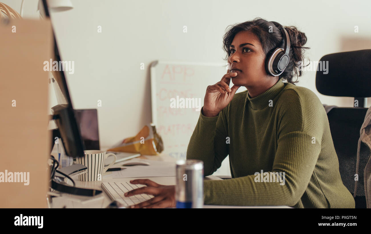 Young woman working on computer in tech startup office. Female programmer sitting at desk and looking away thinking. Stock Photo