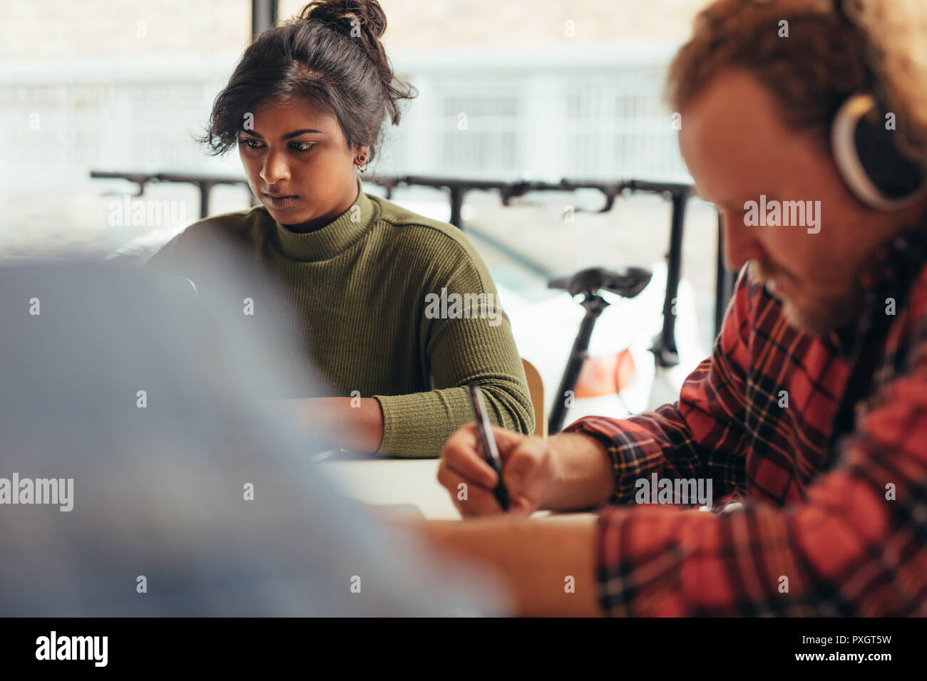 Young woman working on laptop with male colleague sitting in front writing some notes. Computer programmers working at tech startup. Stock Photo