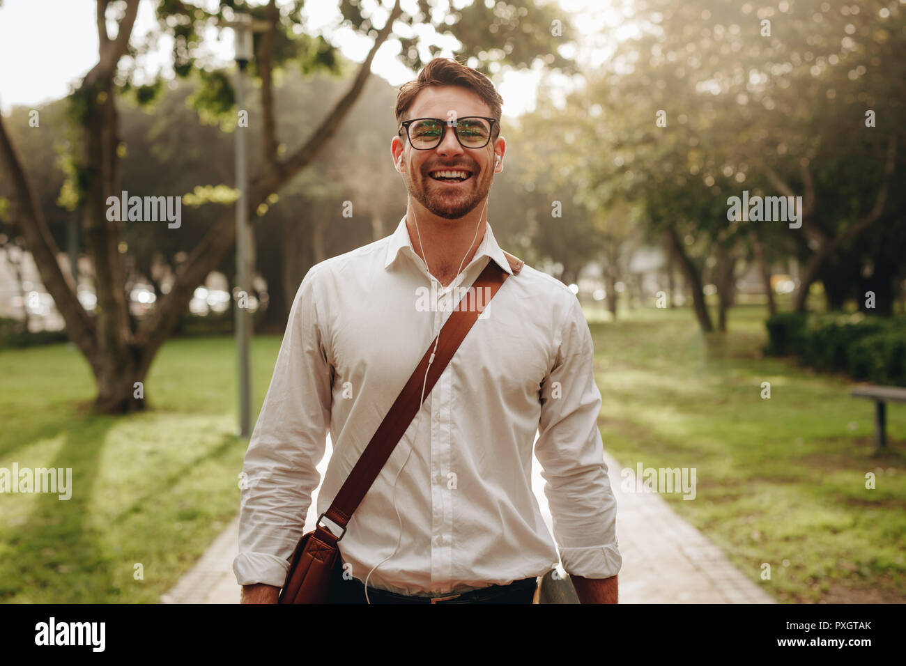 Man walking to office hanging an office bag on his shoulder. Smiling businessman going to office listening to music using earphones. Stock Photo