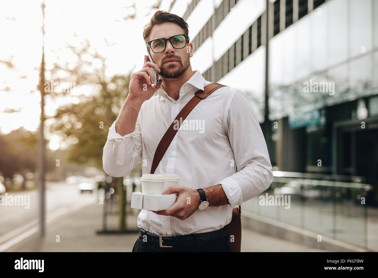 Businessman walking to office talking on mobile phone. Man carrying two coffee cups in hand talking on mobile phone going to office in the morning. Stock Photo