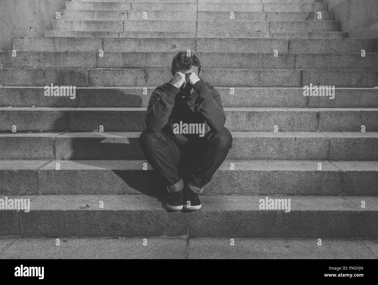 Young desperate jobless man in casual clothes abandoned lost in depression sitting on ground concrete stairs alone in grunge lighting in Emotional pai Stock Photo